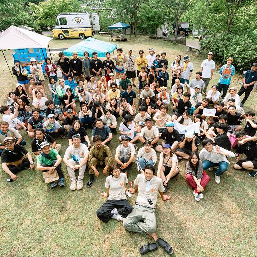 芝生の上で大勢の人が座って記念写真を撮っている様子。奥には黄色と白色のカレーのキャンピングカーが見える。